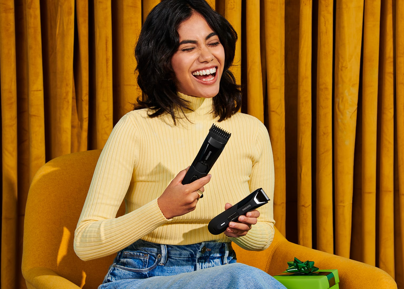 Female model sitting in a chair holding The Beard Hedger in her right hand and The Lawn Mower 5.0 Ultra in her left hand looking happy
